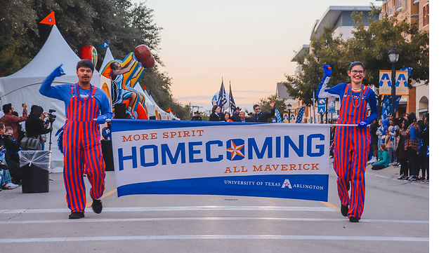Two ambassadors in blue and orange stripes walking down the street with a sign between them labelled 'HOMECOMING'.