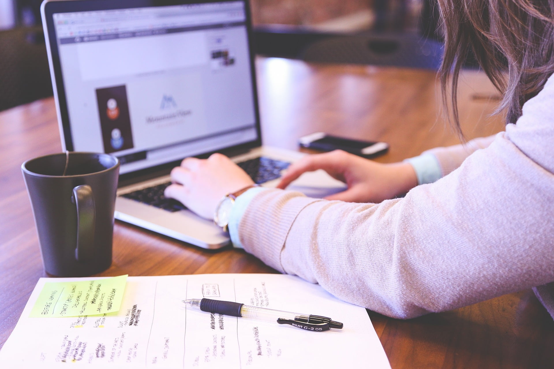 Student sitting at a table with a coffee mug and a notebook, scrolling on a laptop.